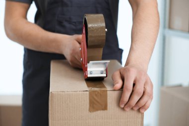 Karton kutuyu kapalı bir yere kapatmak için yapıştırıcılı bant kullanan bir adam.