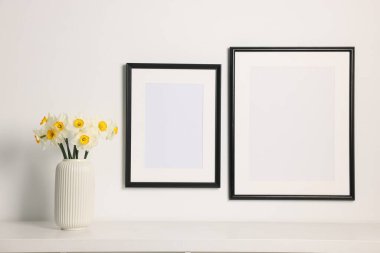 Beyaz masadaki vazodaki güzel nergisler ve kapalı alandaki resim çerçeveleri.
