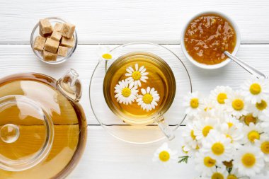 Cam fincanda nefis papatya çayı, kahverengi şeker, çaydanlık, reçel ve taze çiçekler beyaz ahşap masada, düz uzanmış