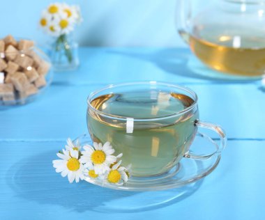 Cam bardakta nefis papatya çayı, taze çiçekler ve açık mavi ahşap masada kahverengi şeker, yakın plan.