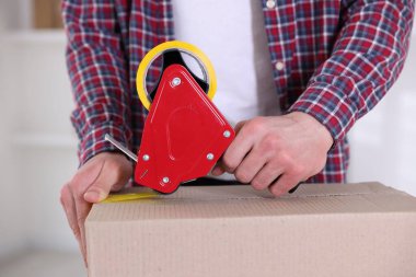 Karton kutuyu kapalı bir yere kapatmak için yapıştırıcılı bant kullanan bir adam.