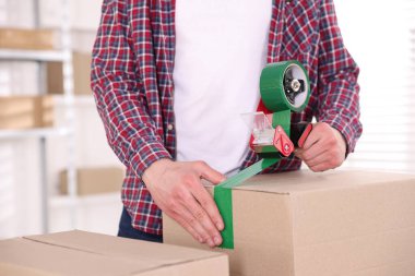 Karton kutuyu kapalı bir yere kapatmak için yapıştırıcılı bant kullanan bir adam.