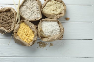 Beyaz ahşap masa üzerinde kağıt torbalarda farklı türde un, düz yerde.