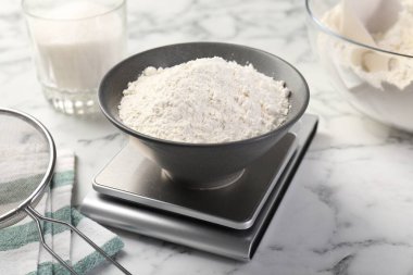 Dijital mutfak terazisi, beyaz mermer masa üzerinde bir kase un ve diğer ürünler, yakın plan.