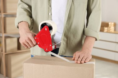 Karton kutuyu kapalı bir yere kapatmak için yapıştırıcılı bant kullanan bir adam.