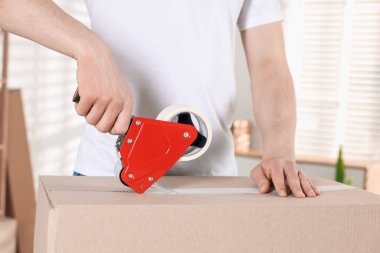 Karton kutuyu kapalı bir yere kapatmak için yapıştırıcılı bant kullanan bir adam.