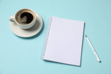Açık mavi arkaplanda defter, kalem ve kahve var.