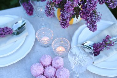 Güzel leylak çiçekleri, zephyrler, tabaklar, çatal bıçaklar, bardaklar ve yanan mumlar bahçede, yukarıdaki manzaranın üzerinde.