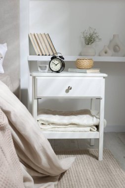 Alarm saati, bir bardak su, hasır kutu ve yatağın yanında kitap olan komodin.