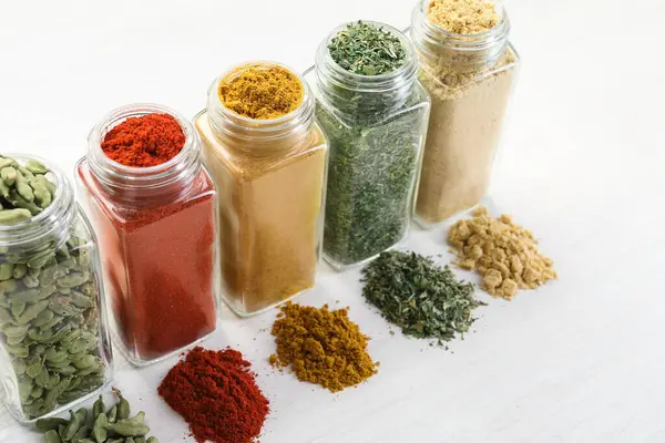 Beyaz masadaki kavanozlarda farklı aromatik baharatlar, yakın plan.