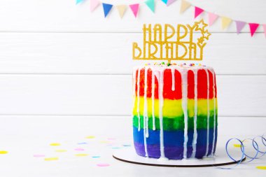 Lezzetli gökkuşağı doğum günü pastası. Masada beyaz ahşap arka planda konfeti ve yazı için yer var.