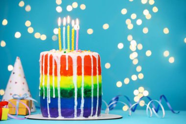 Yakıcı mumlarla, flamalarla, hediye kutularıyla ve açık mavi masada bulanık ışıklara karşı parti şapkasıyla nefis bir gökkuşağı pastası, yakın plan. Metin için boşluk