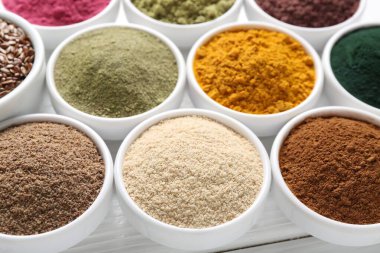 Beyaz ahşap masadaki kaselerde farklı süper yiyecek tozları, yakın plan.