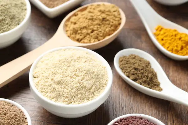 Tabaklardaki farklı süper yiyecek tozları ve tahta masada kaşıklar, yakın plan.