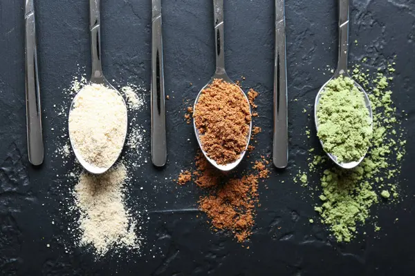 Siyah masanın üzerinde kaşıklarda farklı süper yiyecek tozları var.