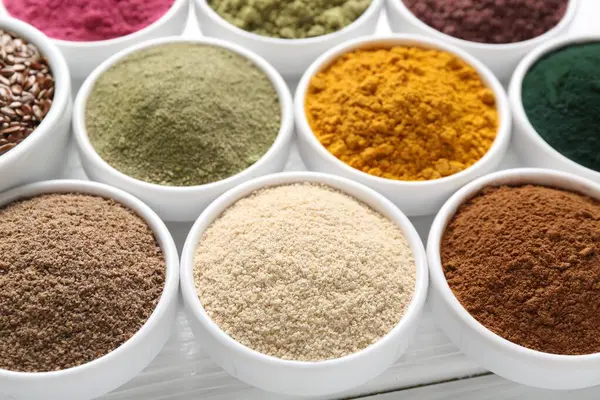 Beyaz ahşap masadaki kaselerde farklı süper yiyecek tozları, yakın plan.