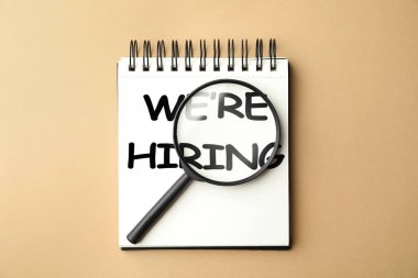 Biz kiralıyoruz. Bej arka planda büyüteç olan defter, üst manzara. İşe alım, insan kaynakları kavramı