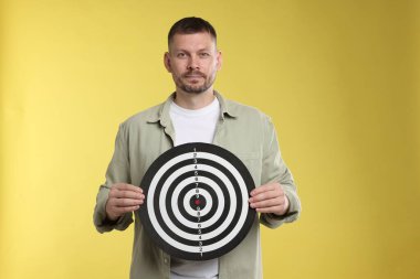 Hedef alınıyor. Sarı arka planda dart tahtası olan adam.