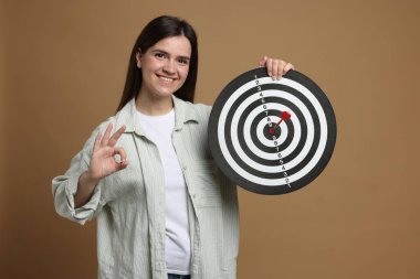 Hedef alınıyor. Dart tahtası ve okla gülümseyen kadın bej arka planda iyi bir jest gösteriyor.