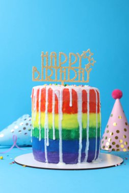 Lezzetli gökkuşağı doğum günü pastası, süslemeler ve açık mavi arka planda parti şapkaları.