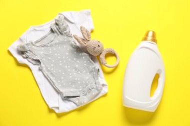 Bir şişe yumuşatıcı, oyuncak ve bebek kıyafetleri sarı arka planda, düz yerde.