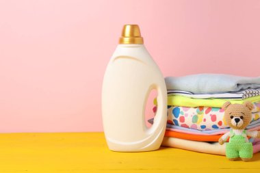 Kumaş yumuşatıcılı şişe, bebek kıyafetleri yığını ve pembe arka planda sarı ahşap masada oyuncak. Metin için boşluk