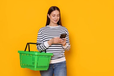 Sarı arka planda akıllı telefonu ve sepeti olan gülümseyen bir kadın. İnternetten alışveriş