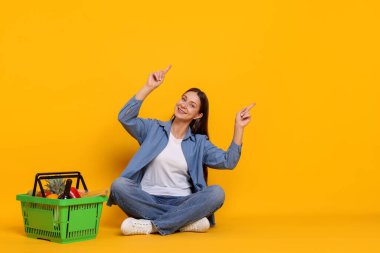 Gülümseyen kadın bir şeyi işaret ediyor ve sarı arka planda ürünlerle dolu bir alışveriş sepeti var. Metin için boşluk