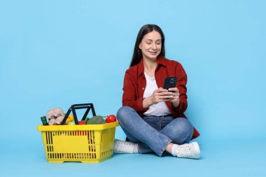 Açık mavi arka planda sepet dolusu ürünleri ve akıllı telefonu olan gülümseyen bir kadın. İnternetten alışveriş