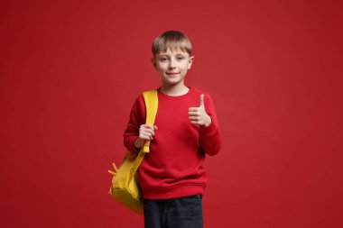 Sırt çantalı yakışıklı okul çocuğu kırmızı arka planda baş parmak gösteriyor.