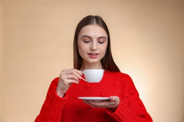 Gülümseyen kadın bej renkli arka planda fincandan kahve içiyor.