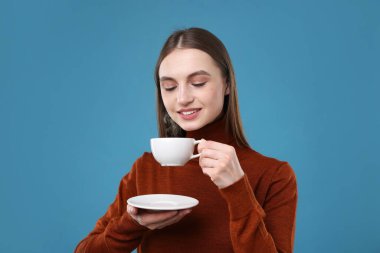 Açık mavi arka planda bir fincan kahveyle gülümseyen kadın.