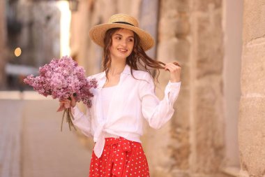 Şapkalı gülümseyen kadın, bina yakınlarında bir demet leylak çiçeğiyle.