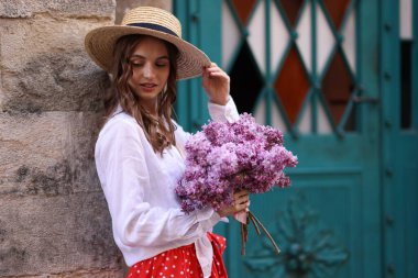Şapkalı güzel bir kadın ve bina yakınlarında bir demet leylak çiçeği.