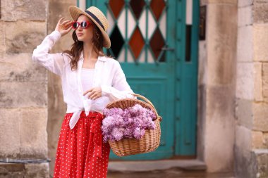 Şapkalı ve güneş gözlüklü güzel bir kadın ve bina yakınlarında bir sepet leylak çiçeği. Metin için boşluk