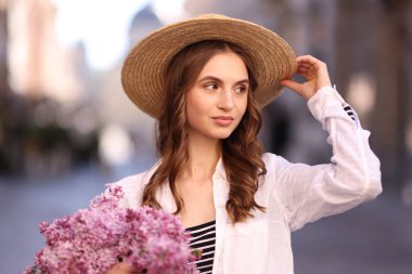 Bulanık arka planda bir demet leylak çiçeğiyle şapkalı güzel bir kadın.