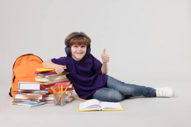 Sırt çantalı, kitaplı ve açık gri arka planda baş parmak gösteren farklı kırtasiyeli sevimli okul çocuğu.
