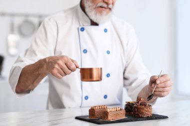 Mutfaktaki masada erimiş çikolatayla lezzetli tatlılar süsleyen yaşlı pasta şefi, yakın plan.