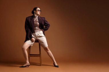 Kahverengi arka planda sandalyede şık giyinmiş genç bir kadın, mesaj için yer var.