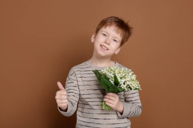 Elinde bir buket güzel çiçekli çocuk kahverengi arka planda baş parmağını gösteriyor.