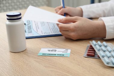 Doktor evrakları hazırlıyor, sağlık sigortası kartı ve haplar ahşap masada, yakın plan.