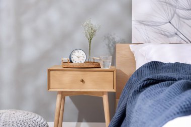Komodinin yanında çalar saat, vazoda çiçekler ve yatağın yanında bir bardak su.