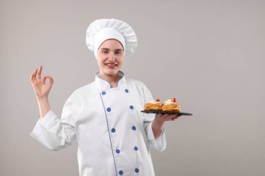 Üniformalı bir şekerci lezzetli profiteroller tutuyor ve açık gri arka planda iyi bir jest gösteriyor.