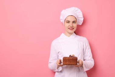 Üniformalı pastacı pembe arka planda çikolatalı kek tutuyor, mesaj için yer var.