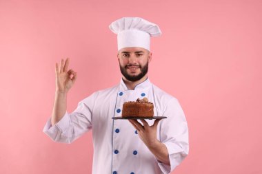 Üniformalı mutlu pastacı elinde çikolatalı pasta ve pembe arka planda güzel bir jest yapıyor.