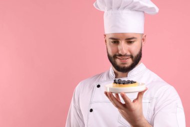Üniformalı mutlu pastacı pembe arka planda yaban mersinli tart tutuyor.