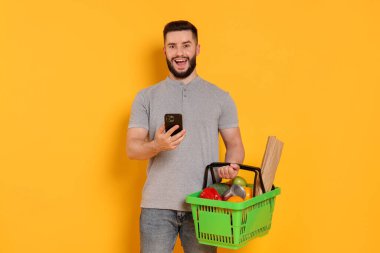 Ürünlerle dolu alışveriş sepeti ve turuncu arka planda akıllı telefonu olan adam.