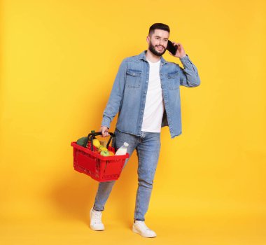 Elinde alışveriş sepeti dolusu mal olan bir adam akıllı telefondan turuncu arka planda konuşuyor.