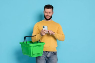 Boş alışveriş sepeti ve açık mavi arka planda akıllı telefonu olan bir adam.