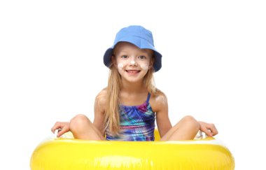 Yüzünde güneş koruyucu kremiyle gülümseyen küçük kız beyaz arka planda şişme halkada oturuyor.